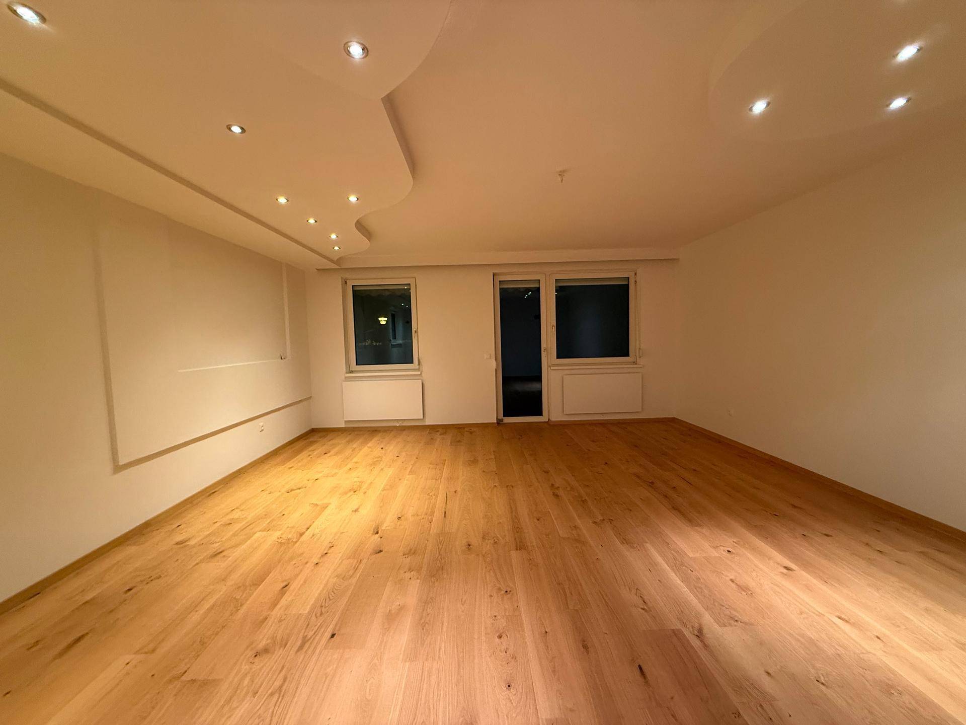 Heller Wohnraum mit elegantem Holzparkett, moderner Deckenbeleuchtung und Zugang zum Außenbereich.