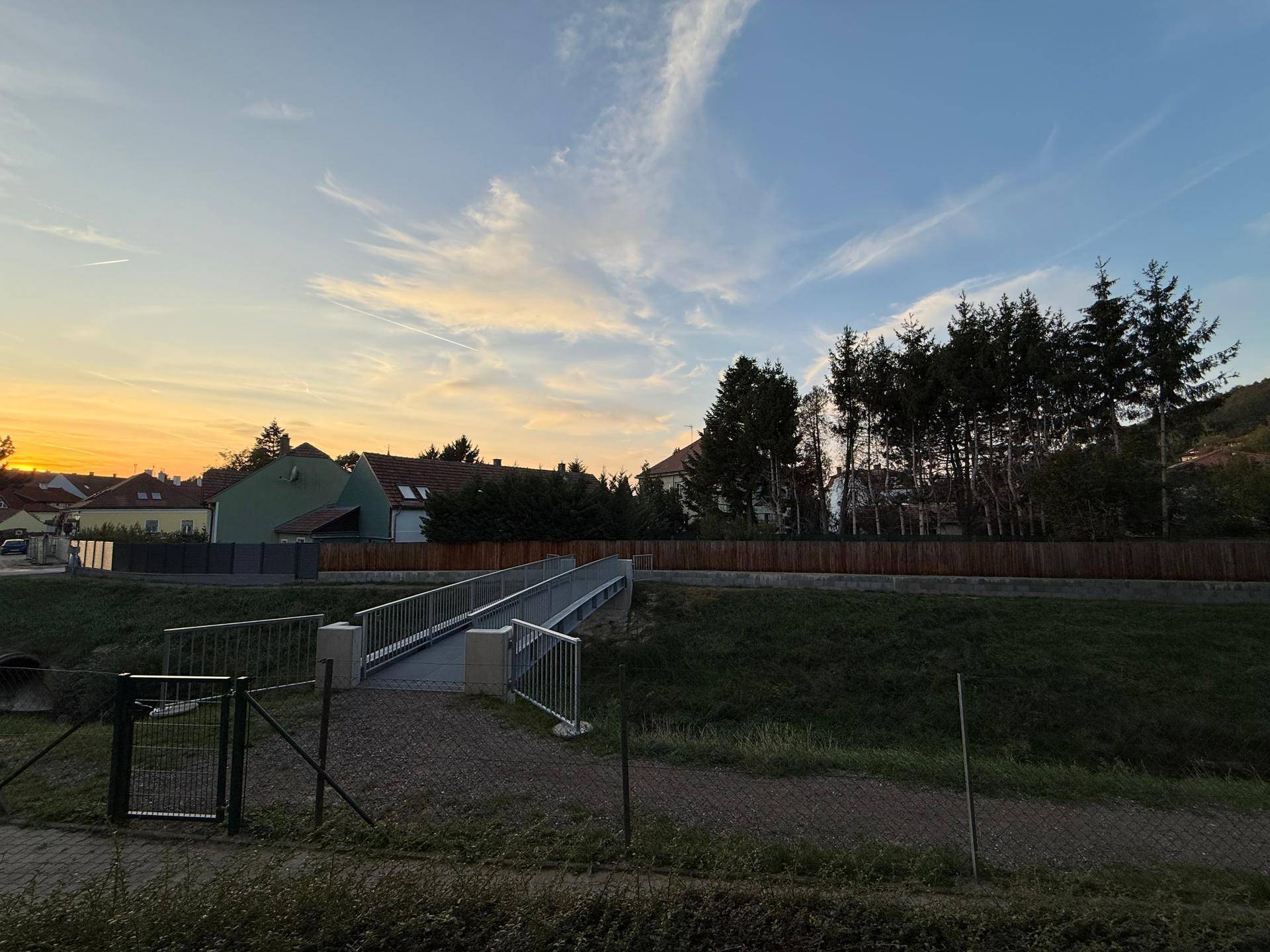 Malerische Außenansicht mit einer Brücke, Bäumen und Häusern im Hintergrund bei Sonnenuntergang.