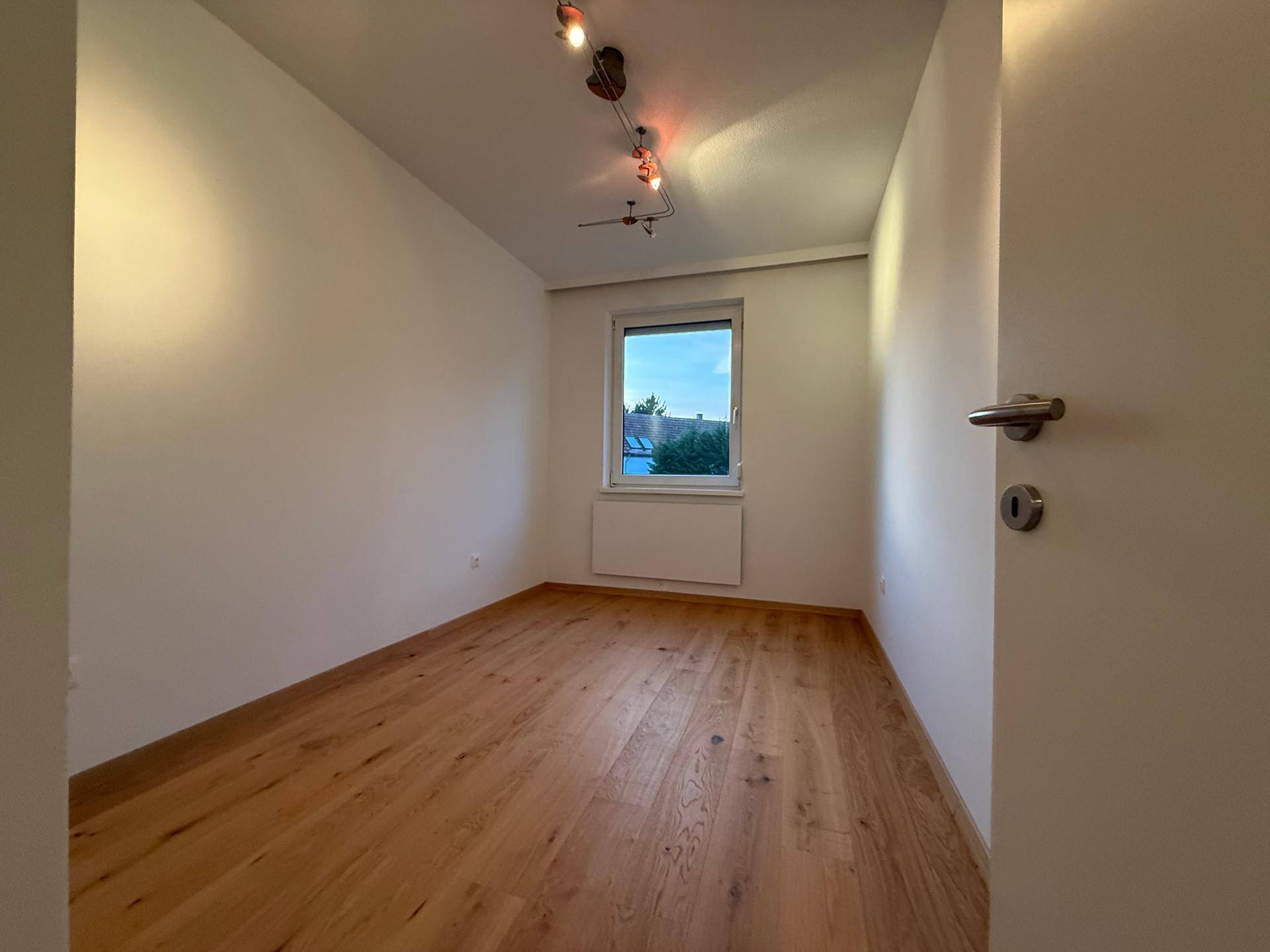 Helles Zimmer mit Holzparkettboden und einem Fenster, das viel Tageslicht hereinlässt.