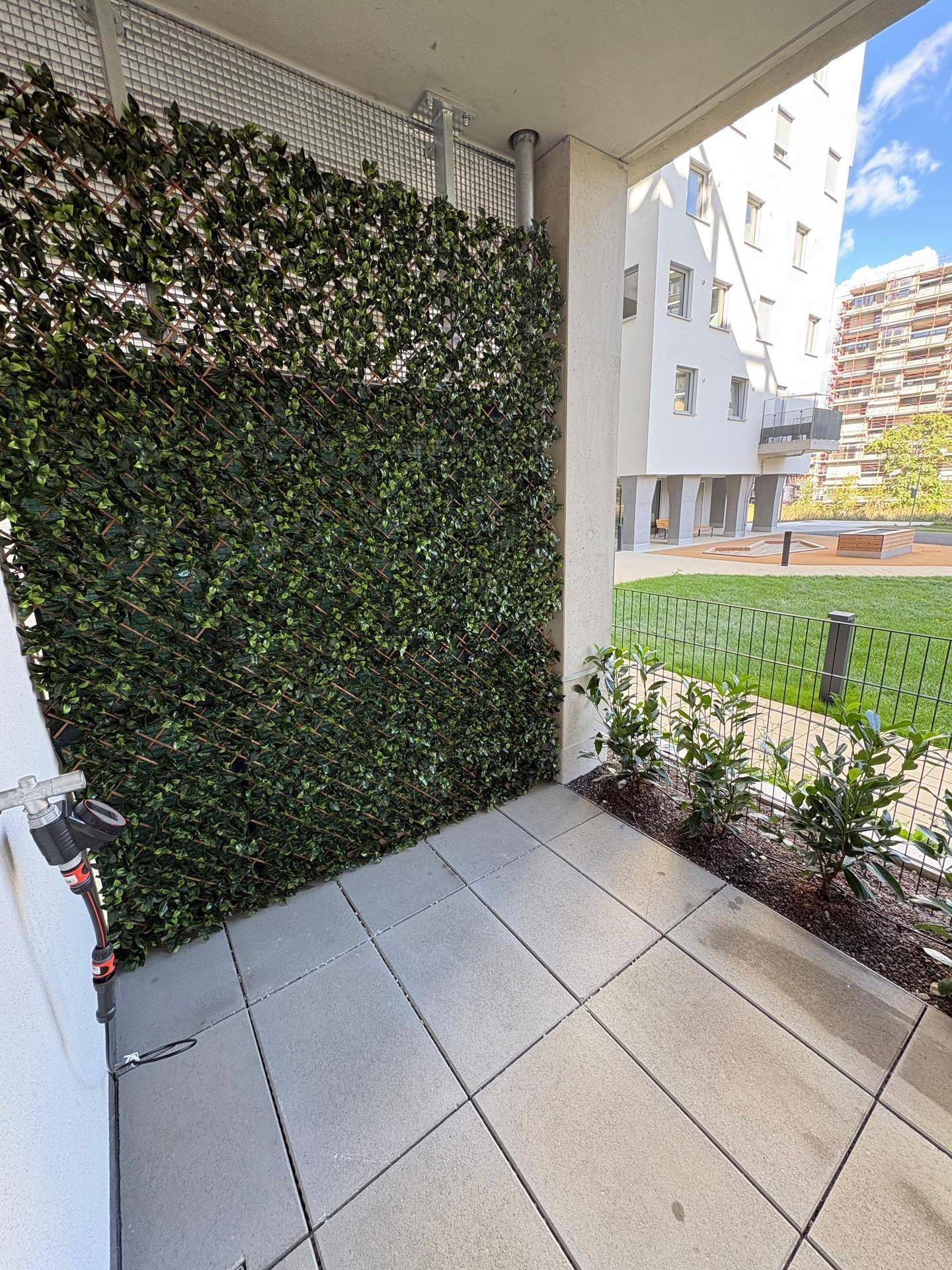 Kleine Terrasse mit Kunstrasenwand, Fliesenboden und Wasseranschluss, angrenzend an den Gemeinschaftsgarten.