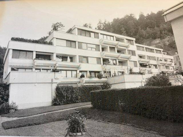 Mehrfamilienhaus mit gestufter Architektur und zahlreichen Balkonen vor bewaldetem Hang.