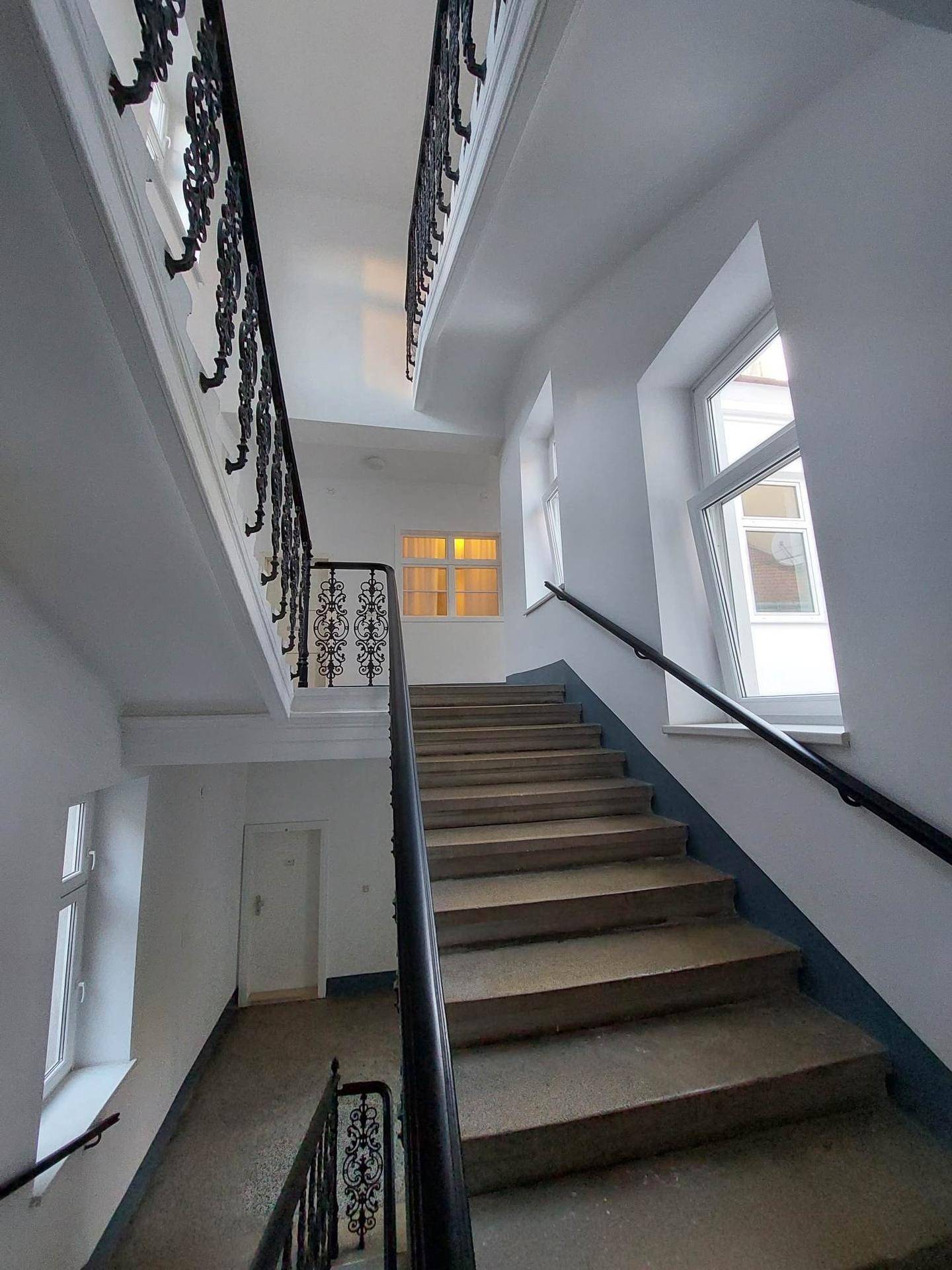 Historisches Treppenhaus mit schmiedeeisernem Geländer und großen Fenstern.