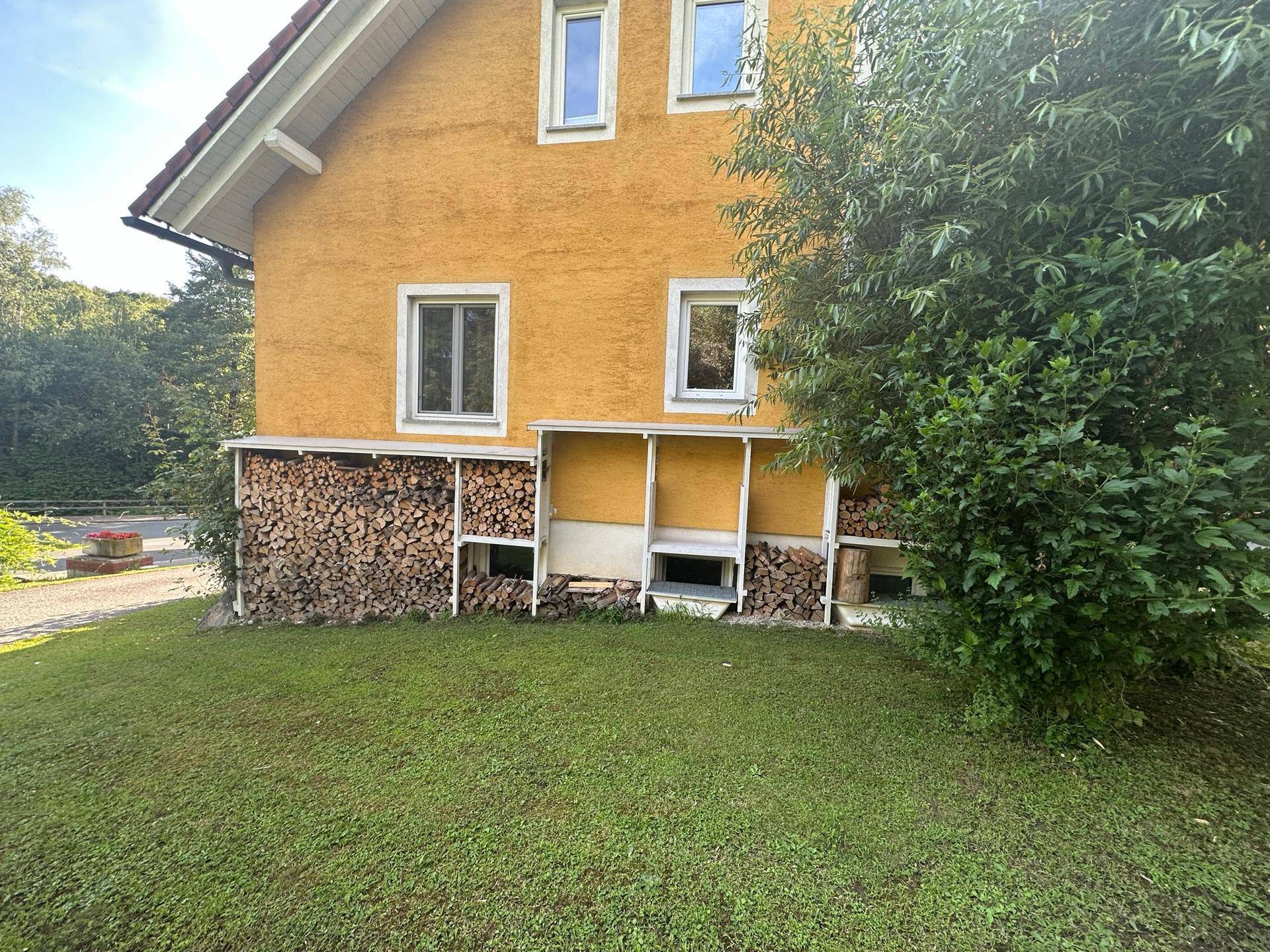 Seitenansicht des Hauses mit gelber Fassade und integriertem Holzstapel.