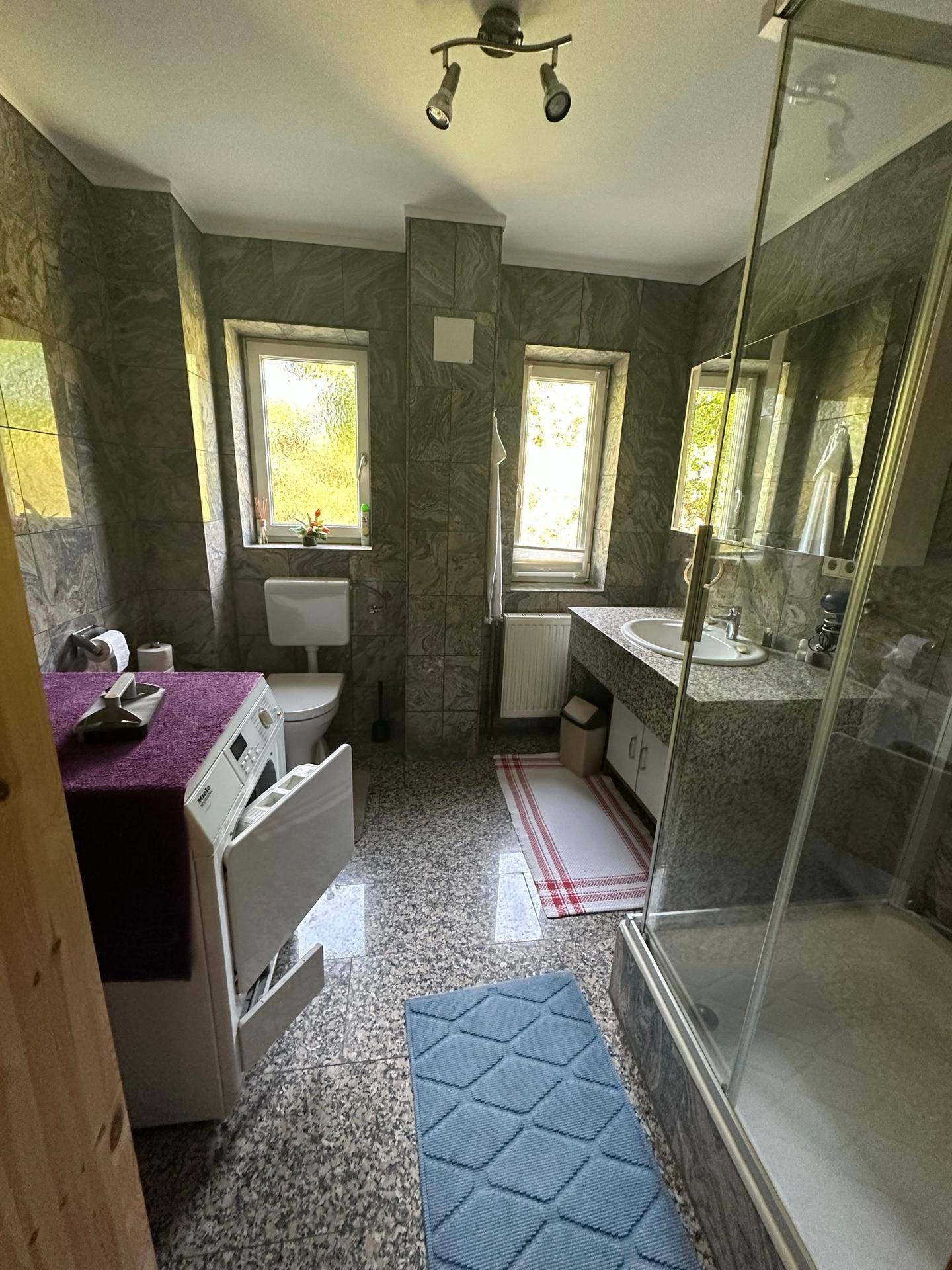 Gefliestes Badezimmer mit Dusche, Waschbecken, Toilette und Waschmaschine.