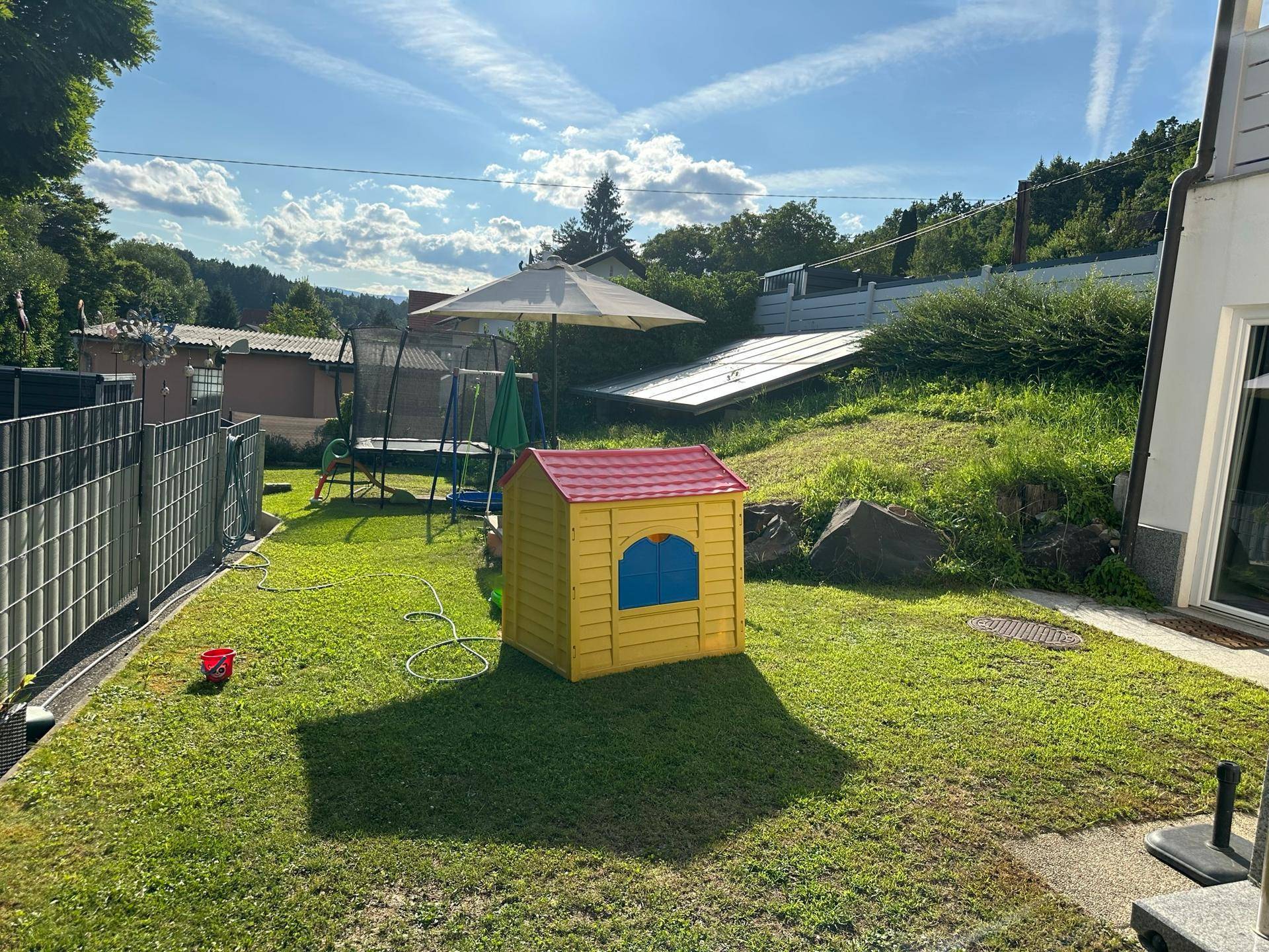 Sonniger Garten mit Rasenfläche, Kinderspielhaus, Trampolin und Photovoltaikanlage im Hintergrund.