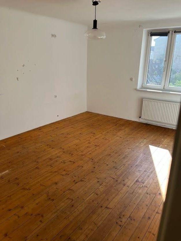Geräumiges Zimmer mit Holzdielenboden, Fenster und Heizkörper, durchflutet von Sonnenlicht.