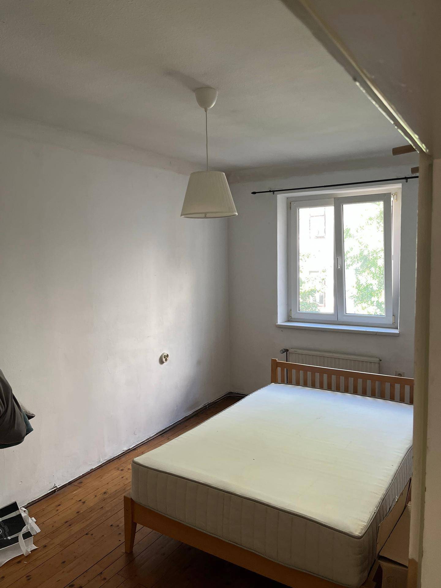 Schlafzimmer mit Holzbett, Dielenboden und Fenster, das natürliches Licht hereinlässt.