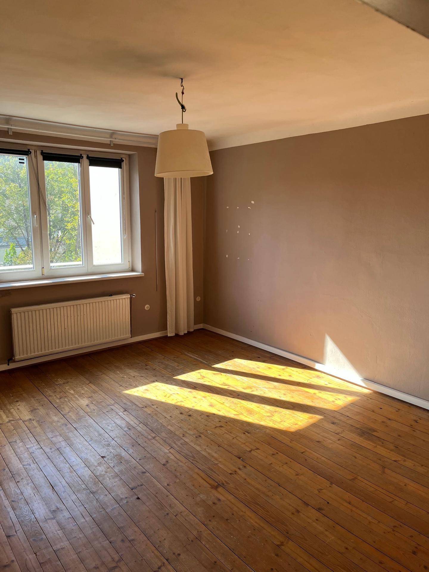 Helles Zimmer mit Holzdielenboden, beigefarbenen Wänden und einem Fenster mit Heizkörper.