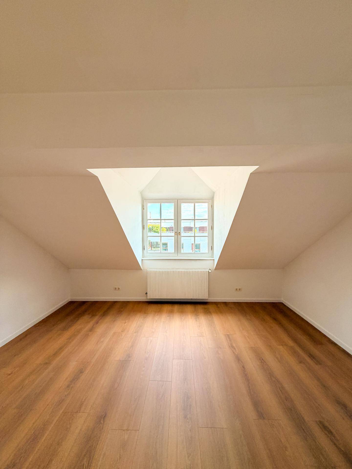 Helles Dachzimmer mit Holzboden, Dachschräge und einem Fenster mit Ausblick.