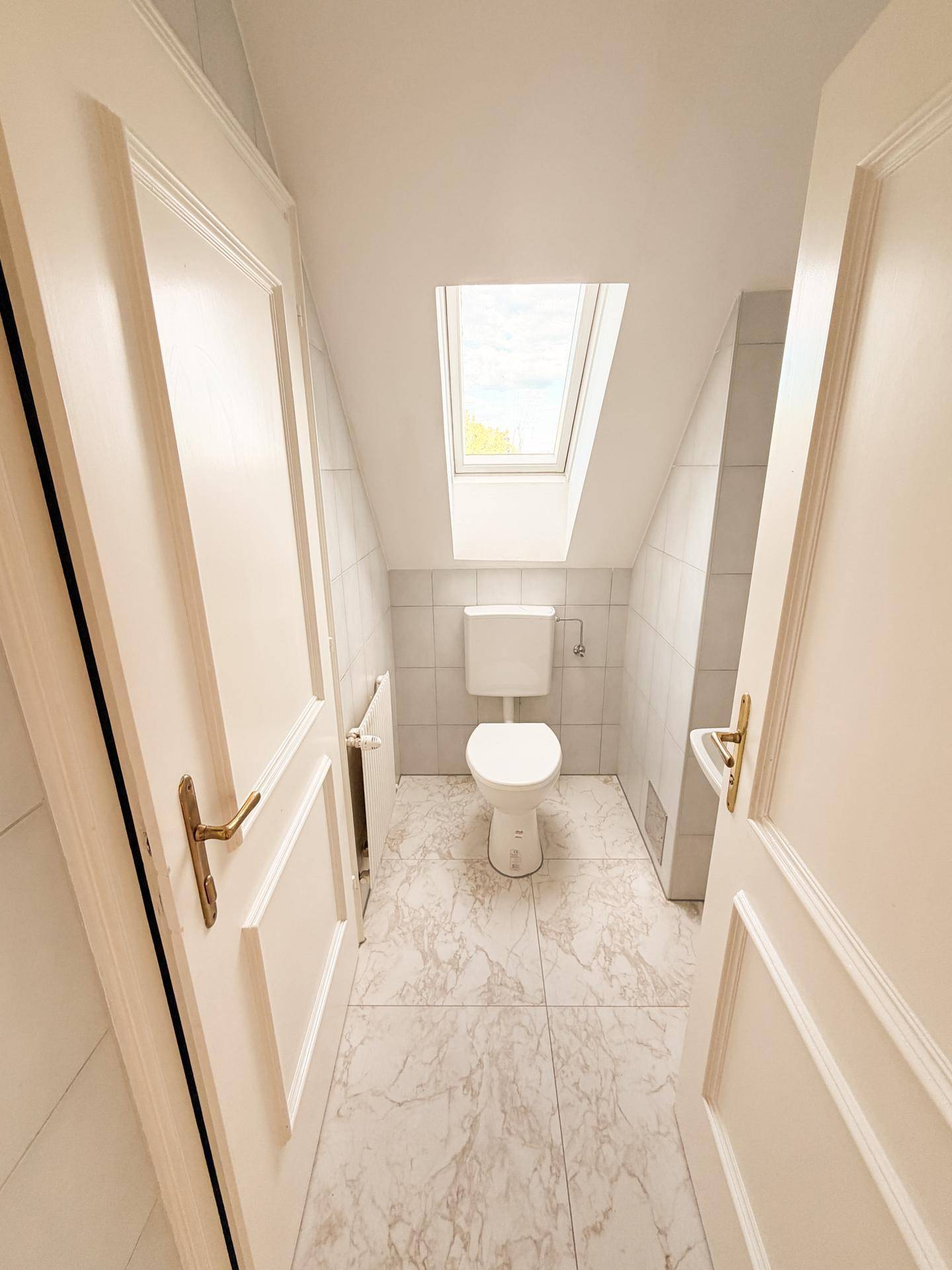 Separates WC mit Fliesenboden, Dachfenster und schrägen Wänden im Dachgeschoss.
