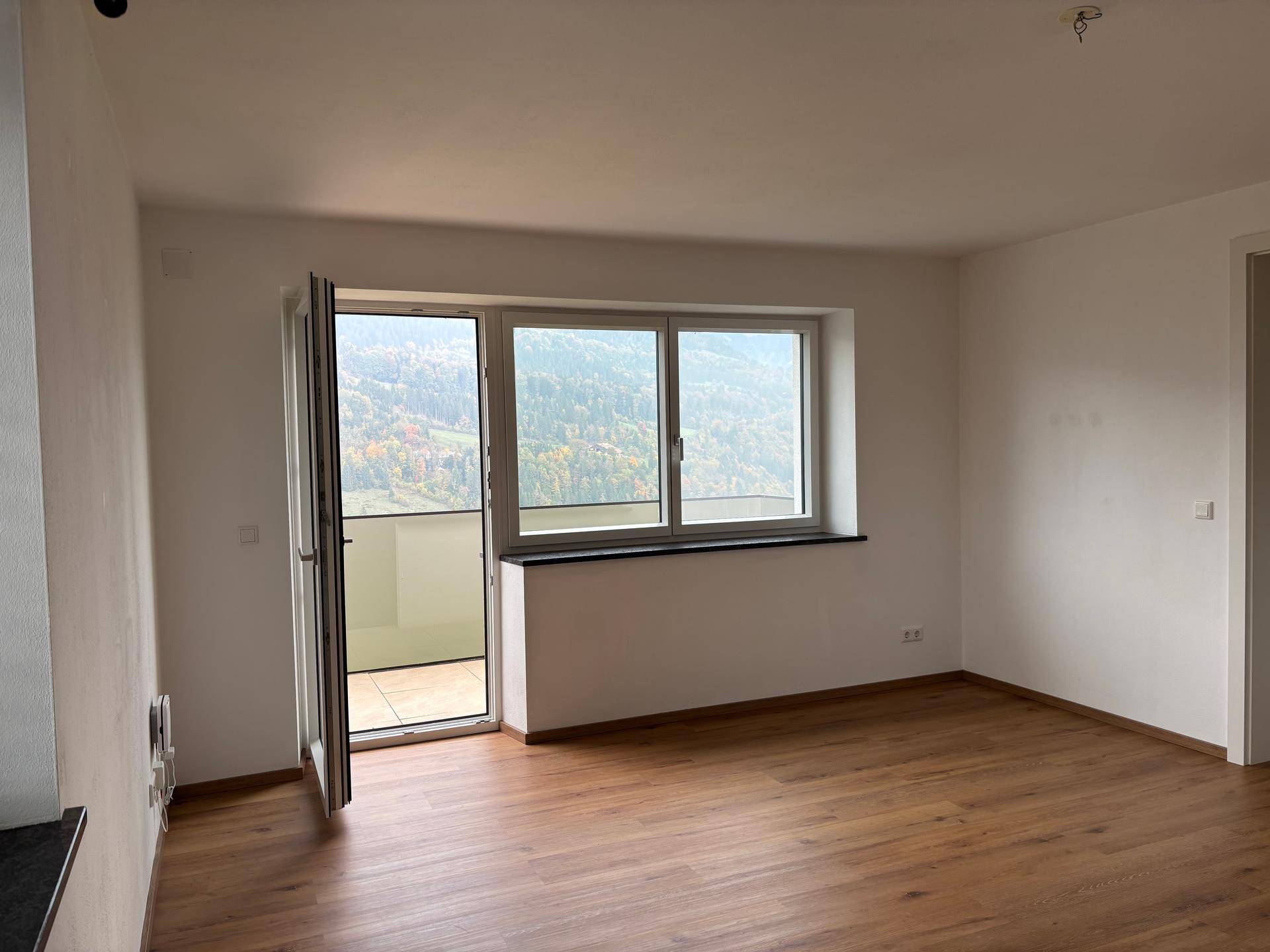 Heller Wohnraum mit Holzboden, Balkonzugang und weitem Blick auf die grüne Landschaft.