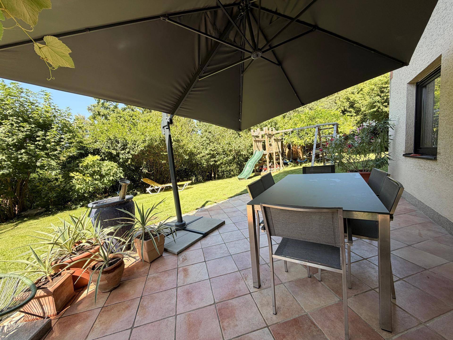 Sonniger Terrassenbereich mit Esstisch, großem Sonnenschirm und Blick auf den Garten mit Spielplatz.