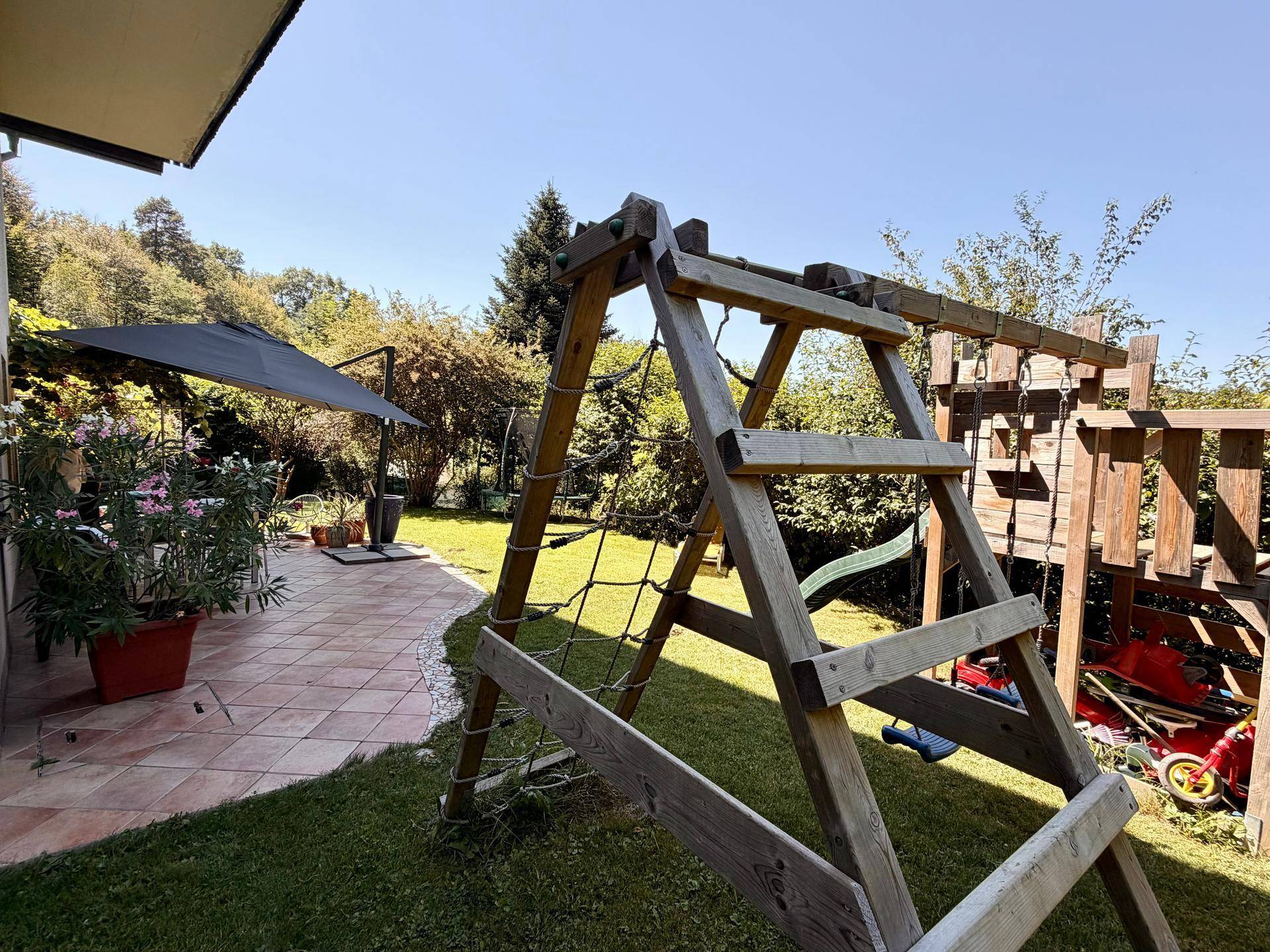 Garten mit Spielplatz und angrenzender Terrasse, umgeben von viel Grün.