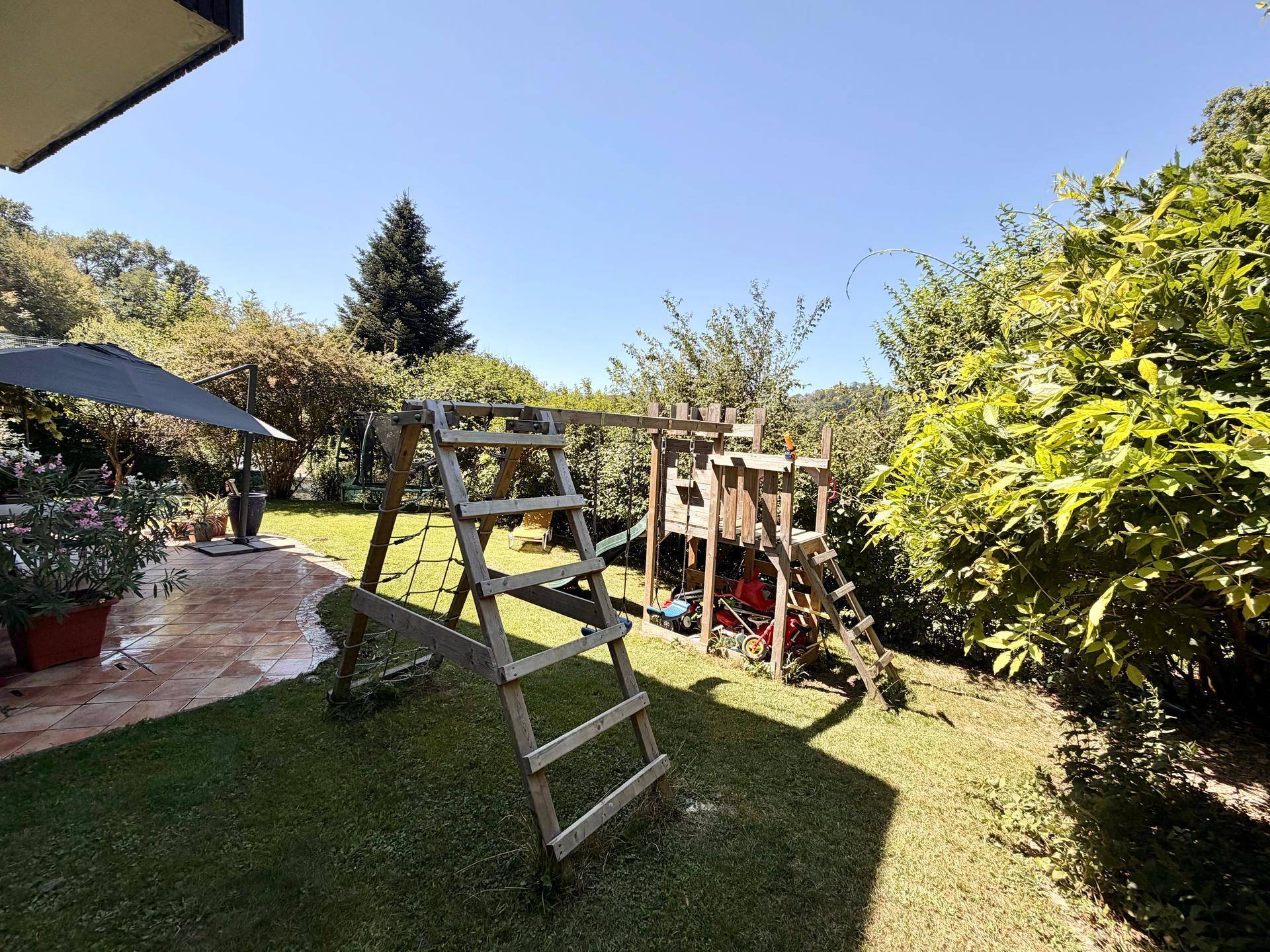 Großer Garten mit Holzspielplatz, vielen Bäumen und Sträuchern unter blauem Himmel.