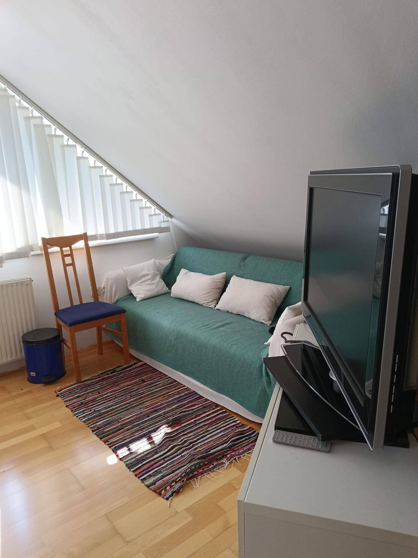 Gemütliches Wohnzimmer mit Sofa, Fernseher und Dachschräge, ausgestattet mit Parkettboden und Jalousien.