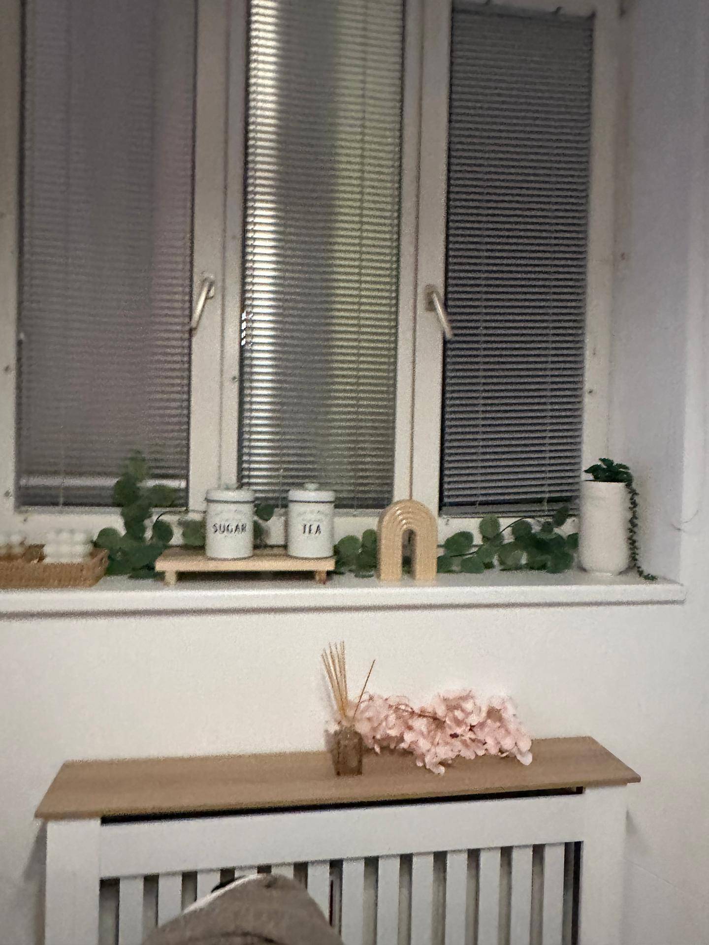 Fenster mit Jalousien und dekorativen Elementen auf der Fensterbank, darunter Teedosen und Blumen.