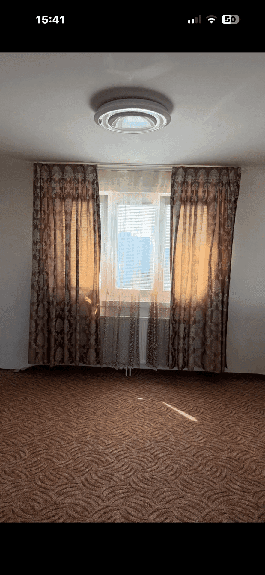 Zimmer mit gemustertem Teppichboden, einem Fenster mit Vorhängen und einer Deckenleuchte.