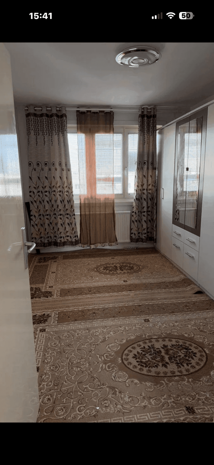 Schlafzimmer mit gemustertem Teppichboden, Spiegelschrank und Fenster mit Vorhängen.
