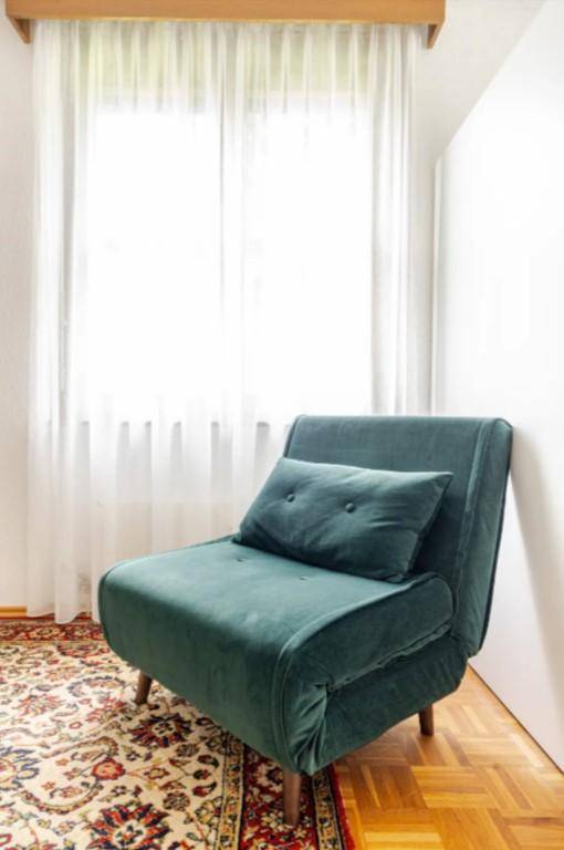 Heller Sitzbereich mit einem grünen Sessel, Teppich und großen Fenstern mit Vorhängen.