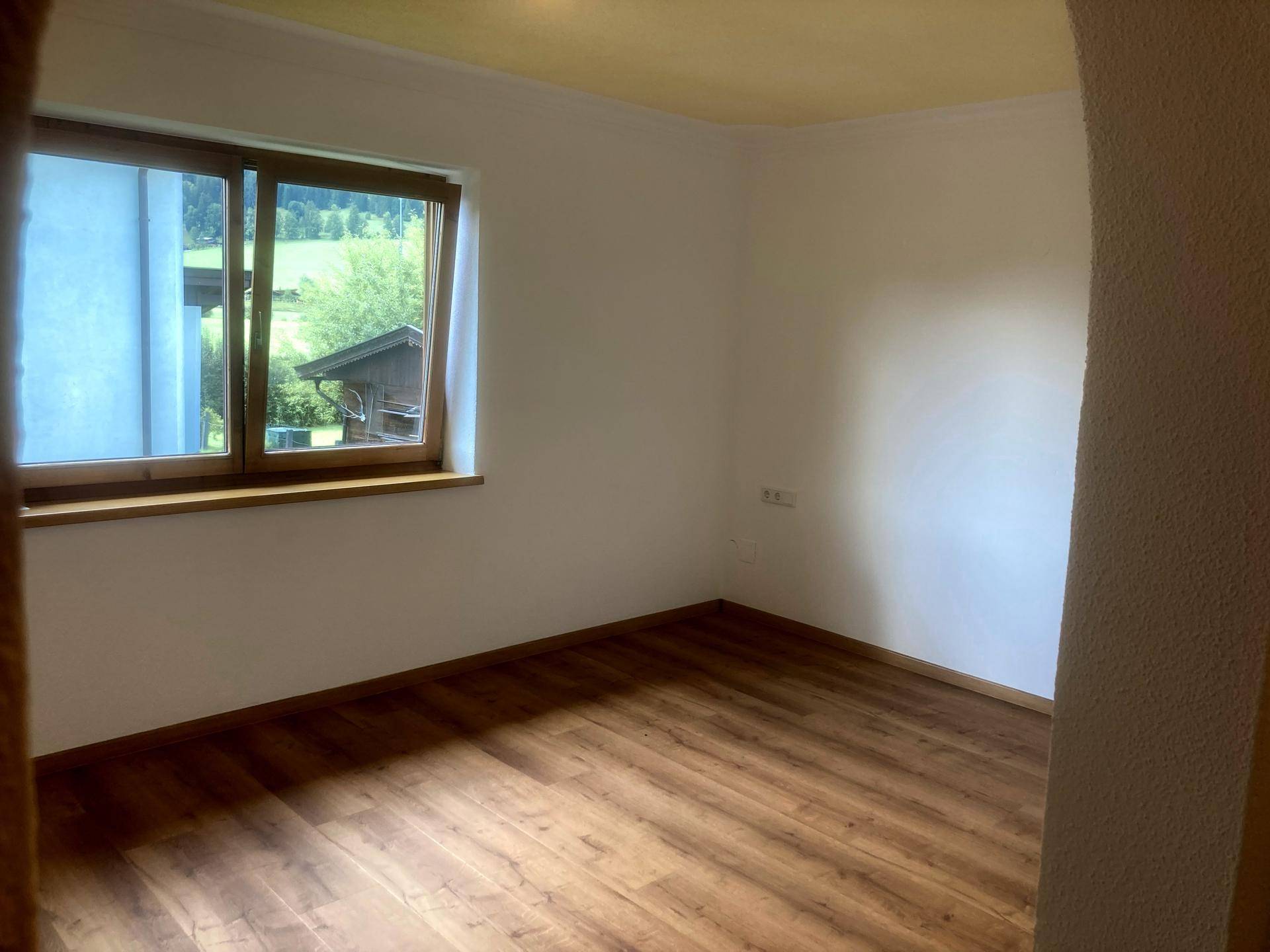 Leeres Zimmer mit Laminatboden und Fenster, das einen ruhigen Blick ins Grüne bietet.