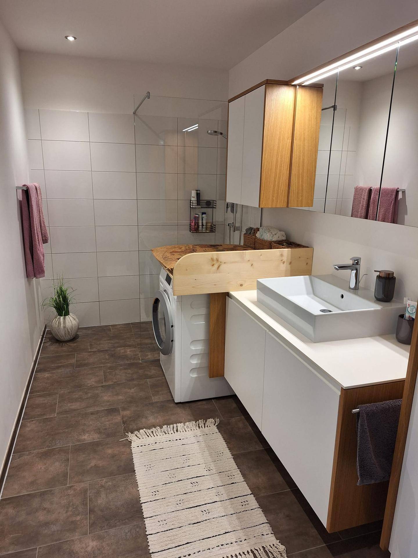 Modernes Badezimmer mit Dusche, Waschbecken und integrierter Waschmaschine unter einer Holzablage.