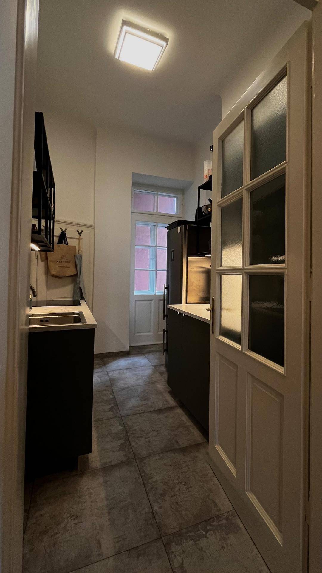 Schmale Küche mit Fliesenboden, schwarzen Schränken und Fenster am Ende des Raumes.
