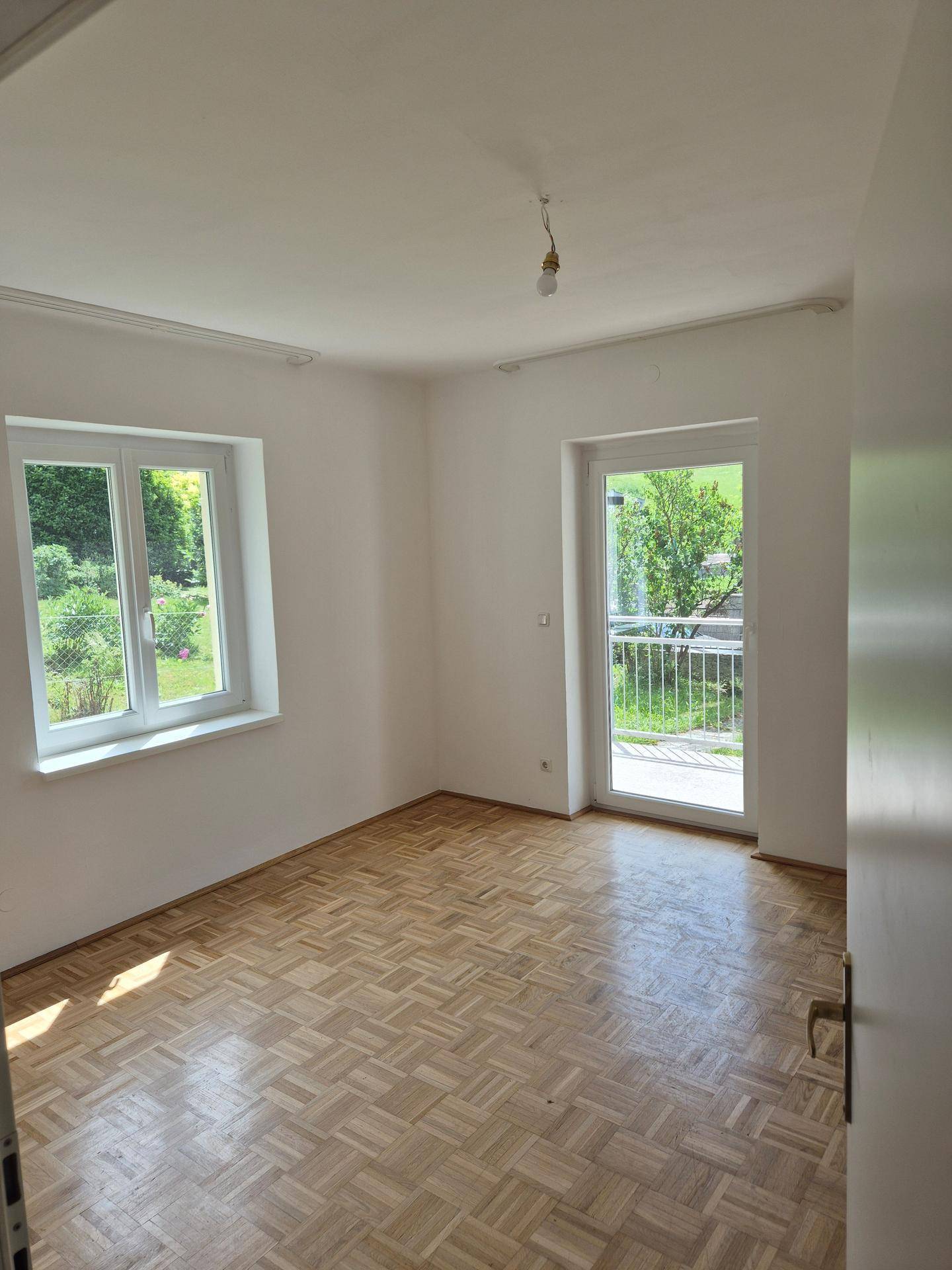 Leeres Zimmer mit Parkettboden, großem Fenster und Zugang zu einem Balkon mit Blick ins Grüne.