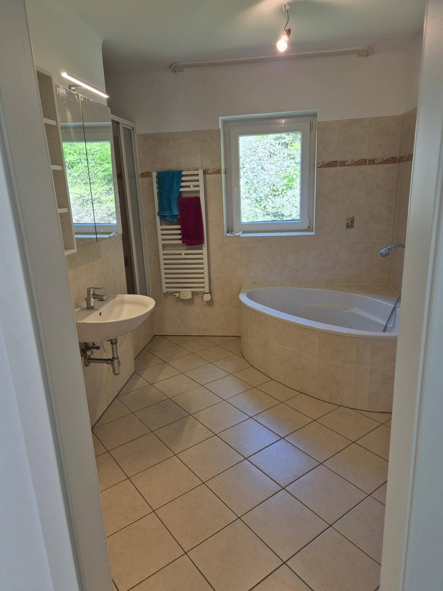 Helles Badezimmer mit Eckbadewanne, separater Dusche, Waschbecken und Fenster für natürliche Belüftung.