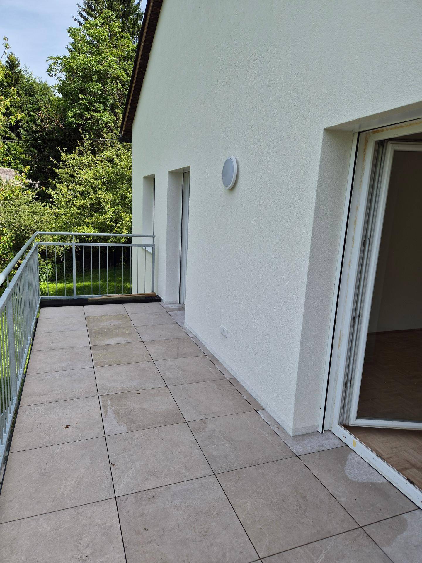 Geräumiger Balkon mit Fliesenboden und Metallgeländer, umgeben von üppigem Grün.