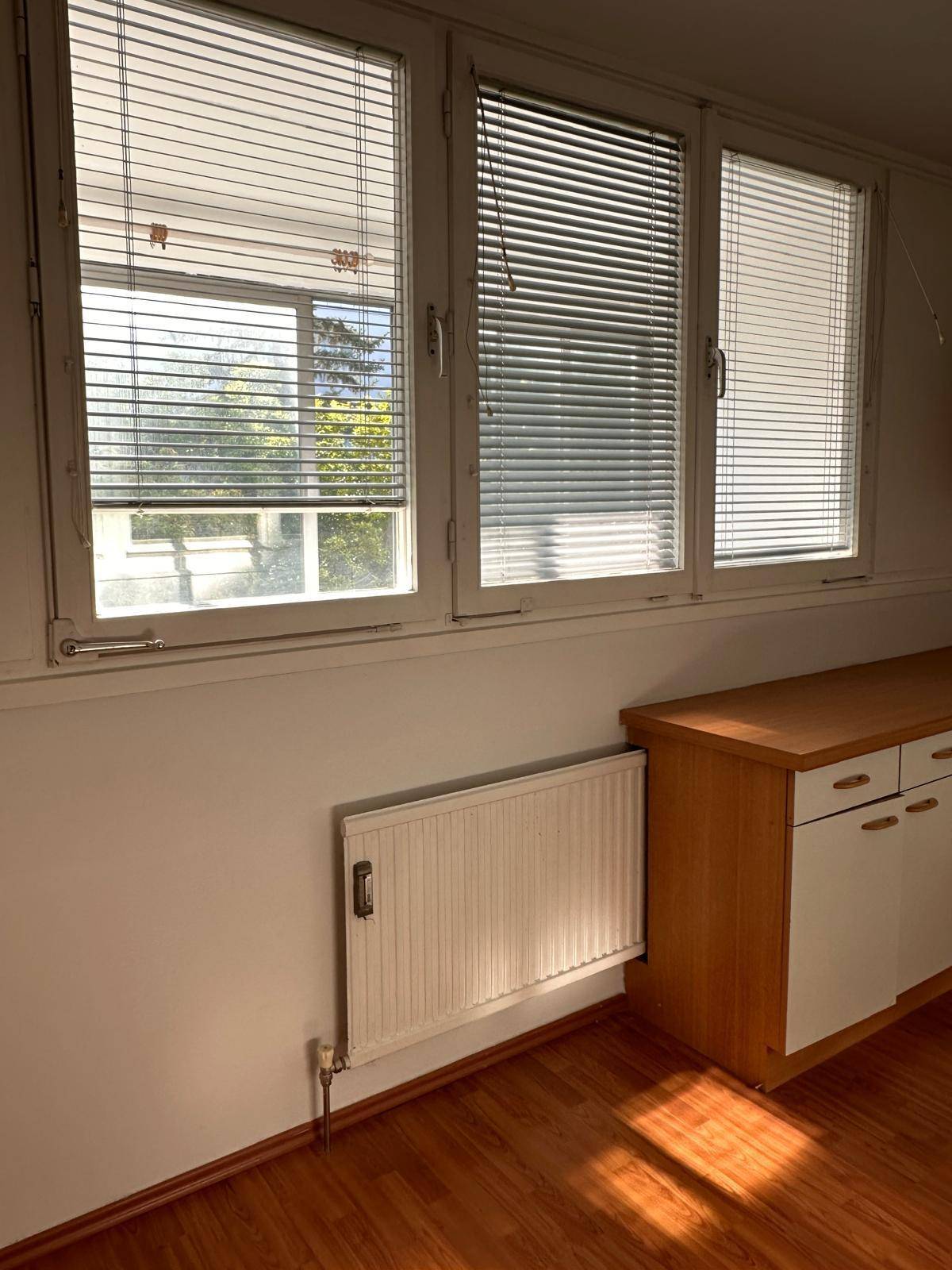 Fensterbereich mit Jalousien, Heizkörper und einem Holzschrank in einem hellen Raum.