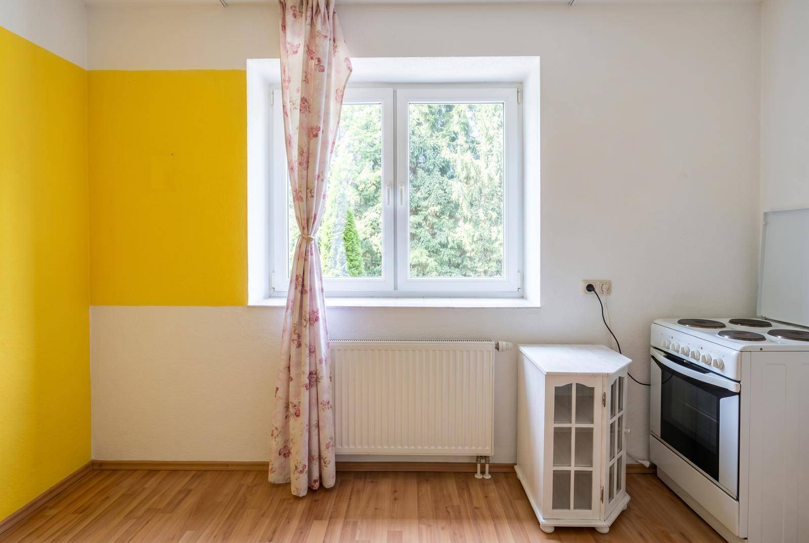 Einfache Küchenzeile mit Herd, Fenster und gelber Wand in einem hellen Raum.