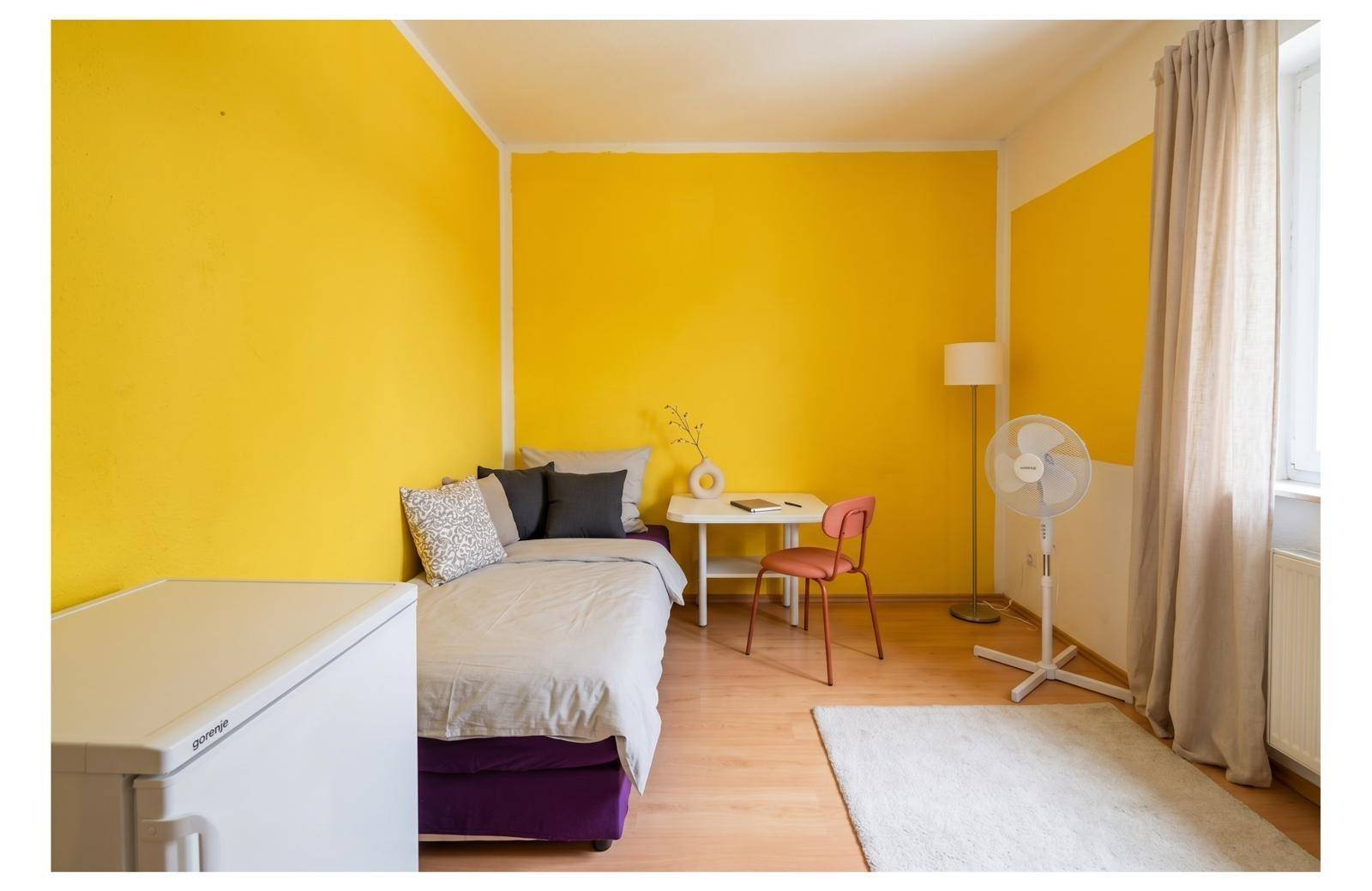 Gemütliches Schlafzimmer mit gelber Akzentwand, Einzelbett und Holzboden.