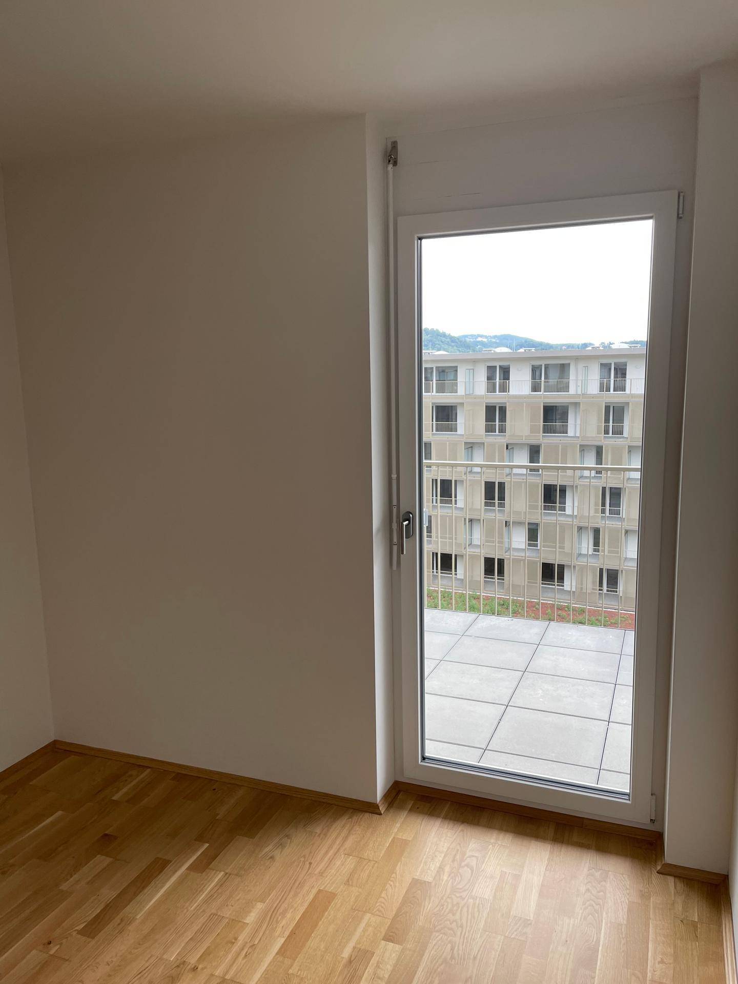 Leeres Zimmer mit Parkettboden und einem großen Fenster, das viel Tageslicht hereinlässt.