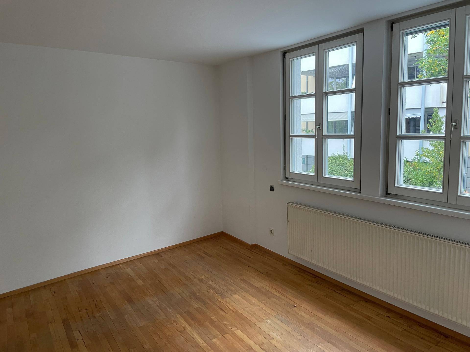 Geräumiges Zimmer mit zwei großen Fenstern, Parkettboden und Heizkörpern, bietet viel Helligkeit.