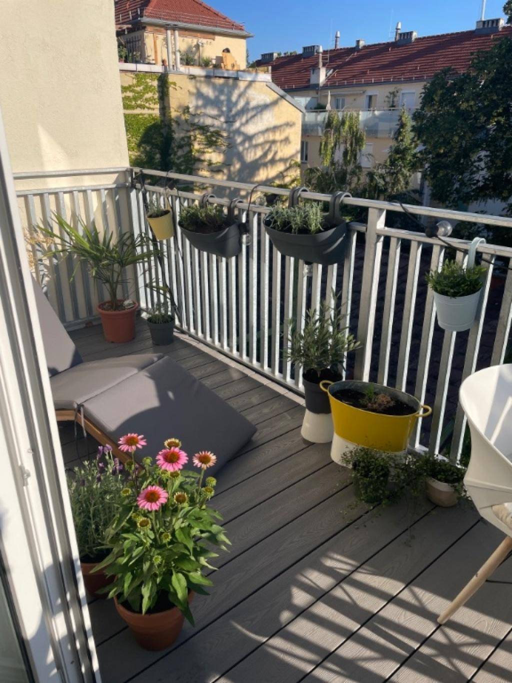 Sonniger Balkon mit Pflanzen, Liegestuhl und Blick auf die umliegenden Gebäude und Dächer.