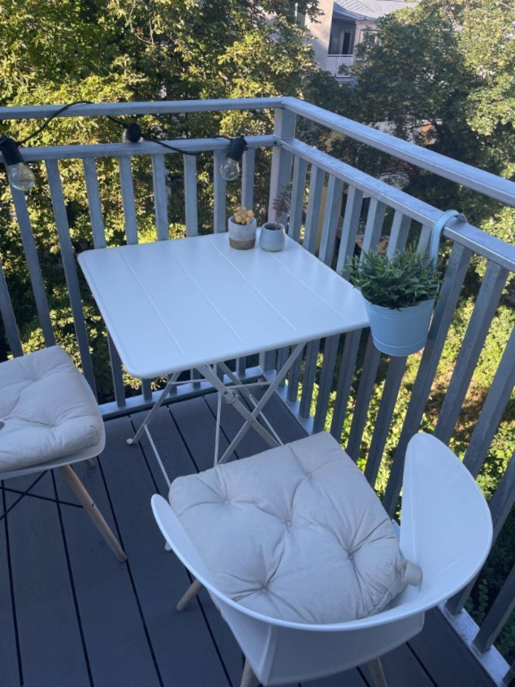 Gemütlicher Balkon mit kleinem Tisch, Stühlen und Lichterkette, umgeben von Bäumen.