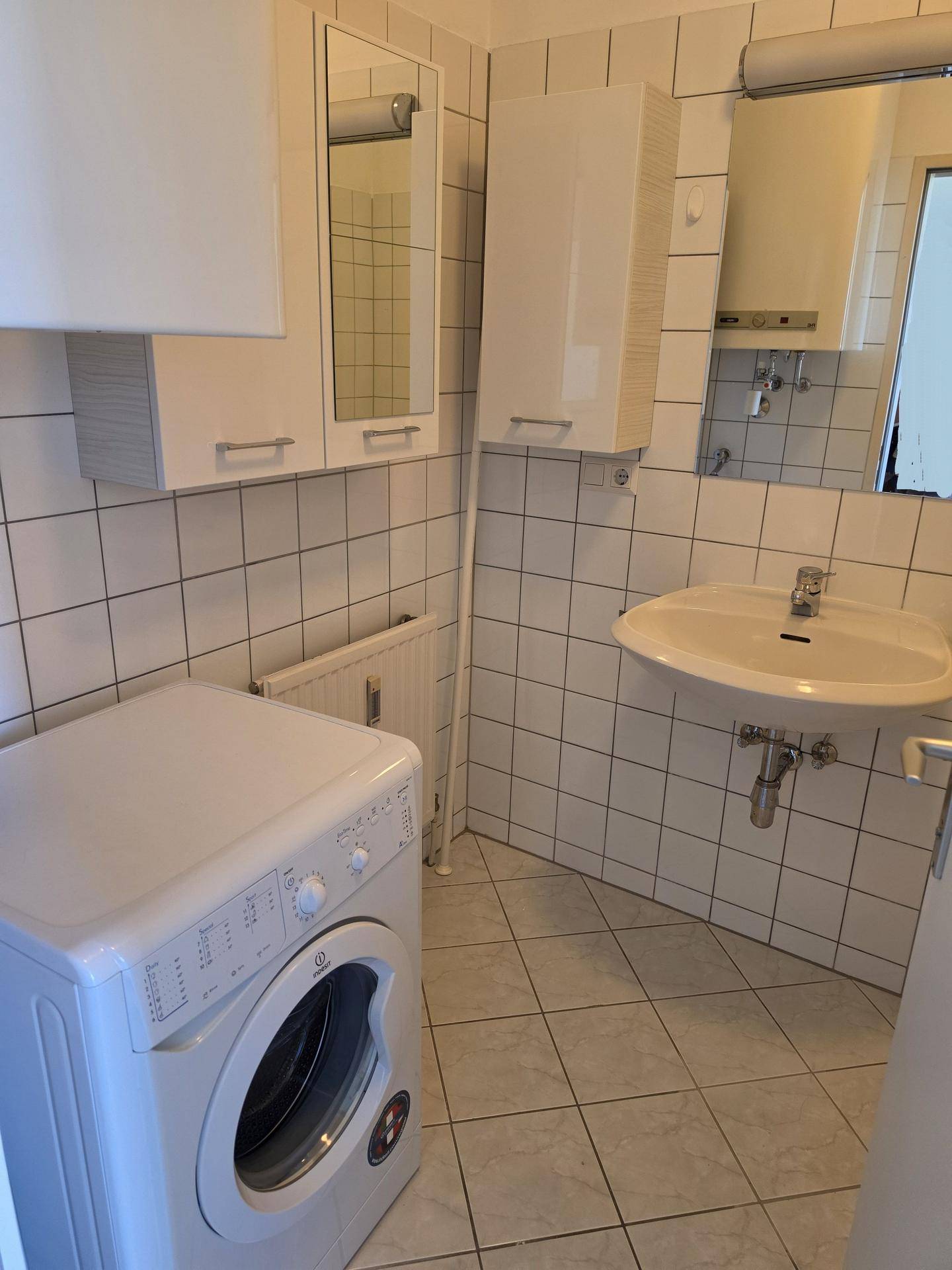 Helles Badezimmer mit Waschbecken, Waschmaschine und weißen Wand- und Bodenfliesen.