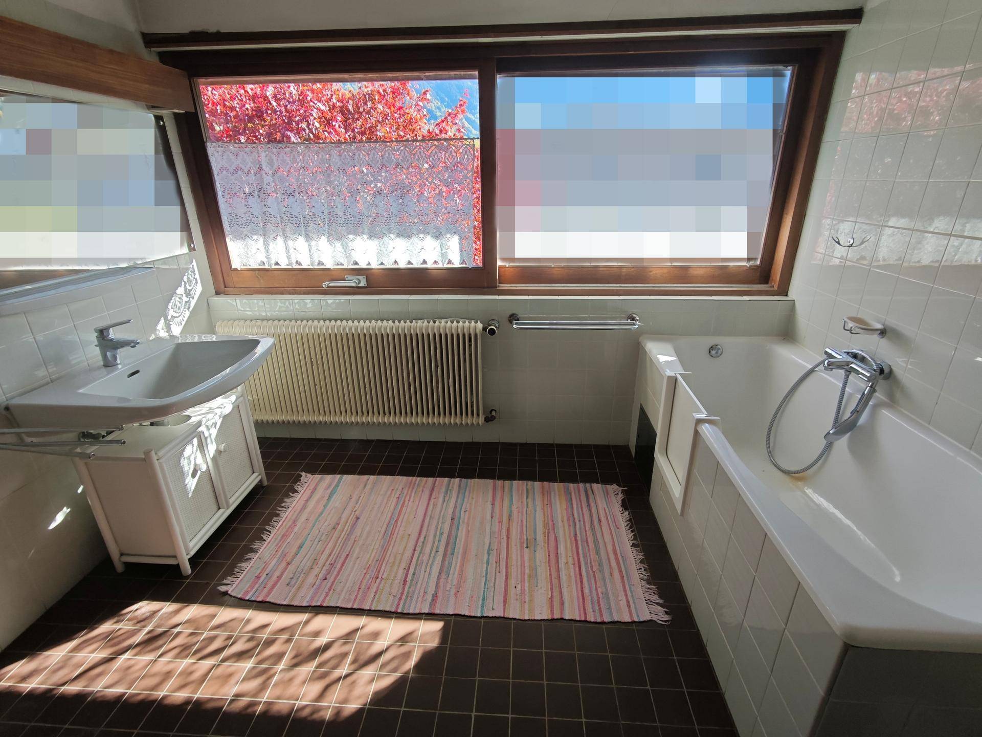 Helles Badezimmer mit Badewanne, Waschbecken, Heizkörper und Fenster, ausgestattet mit weißen Fliesen und braunem Boden.