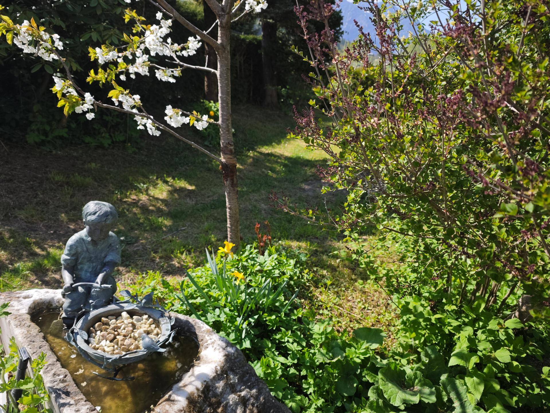 Sonniger Gartenbereich mit blühenden Bäumen, Frühlingsblumen und einer kleinen Steinfigur, die eine idyllische Szene bildet.