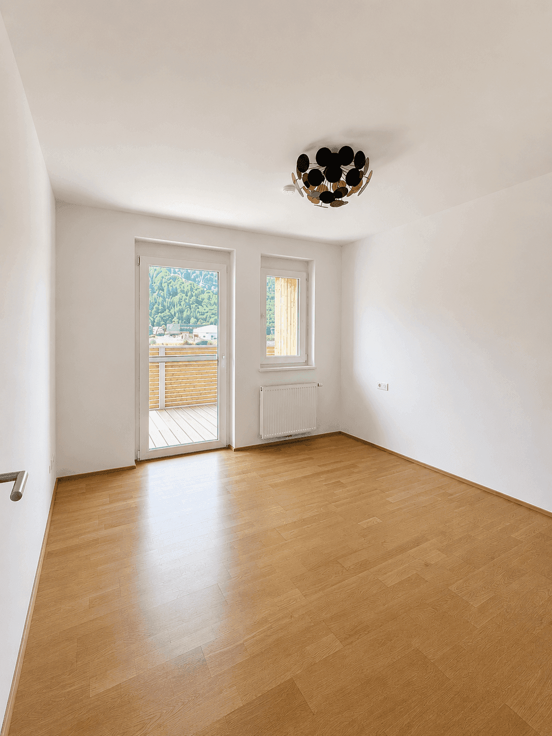 Schlafzimmer mit Holzboden, Heizkörper und Zugang zu einem privaten Balkon mit Holzlattung.