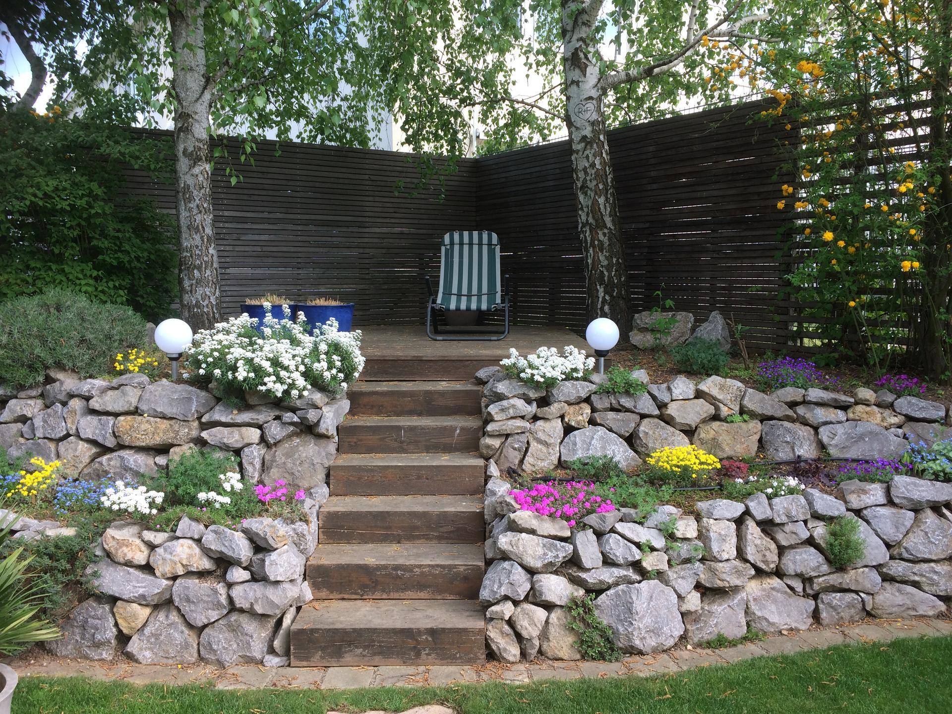 Gestufter Gartenbereich mit Steinmauern, Holztreppe und Sitzgelegenheit unter Birkenbäumen.