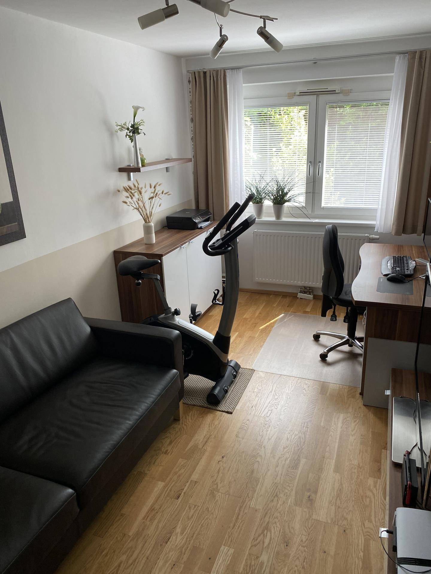 Arbeitszimmer mit Schreibtisch, Sofa und Heimtrainer, gut beleuchtet durch ein Fenster.
