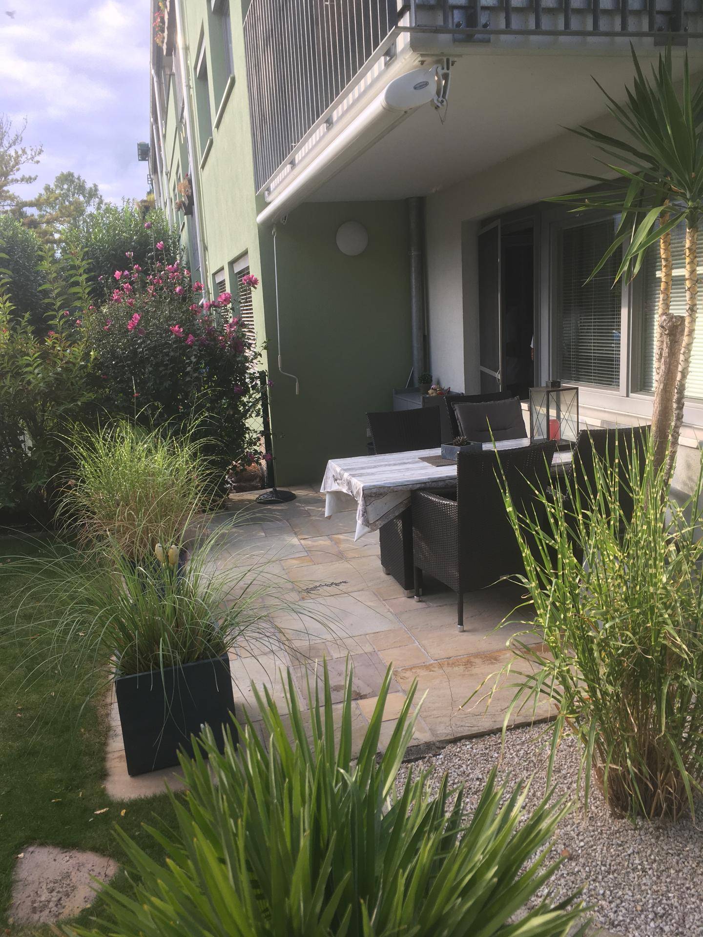Geflieste Terrasse mit Esstisch und Stühlen, umgeben von Pflanzen und einem Balkon darüber.