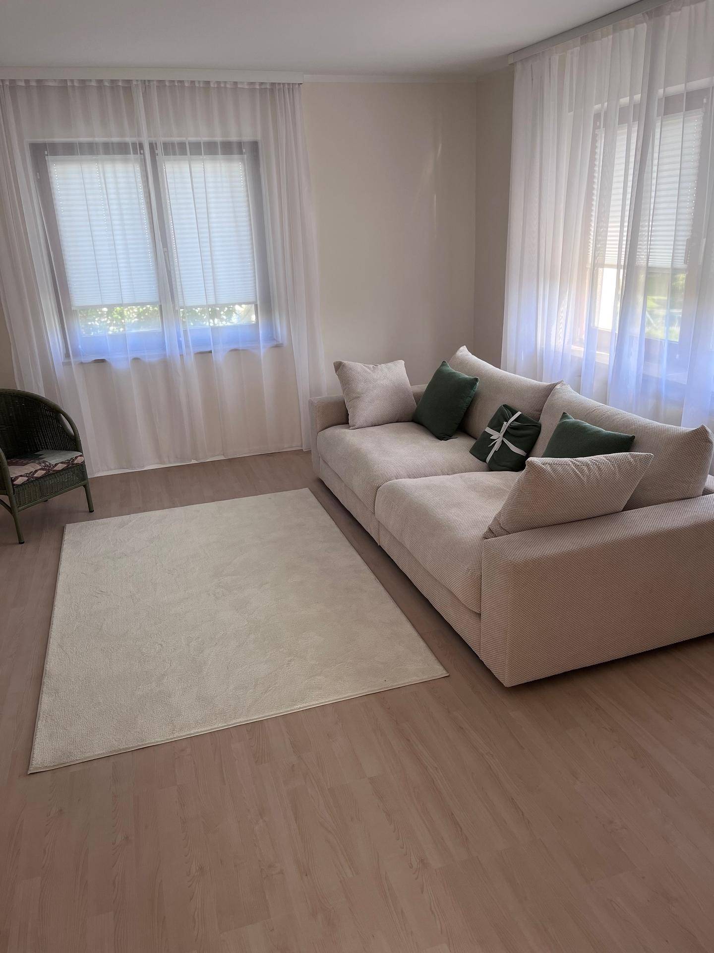 Gemütliches Wohnzimmer mit großem Sofa, hellem Teppich und viel Tageslicht durch die Fenster.