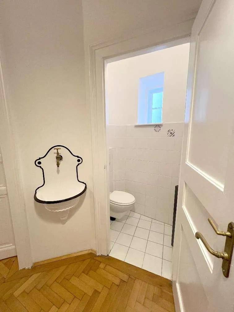 Separate Toilette mit kleinem Waschbecken und Fenster, hell gefliest und sauber gehalten.