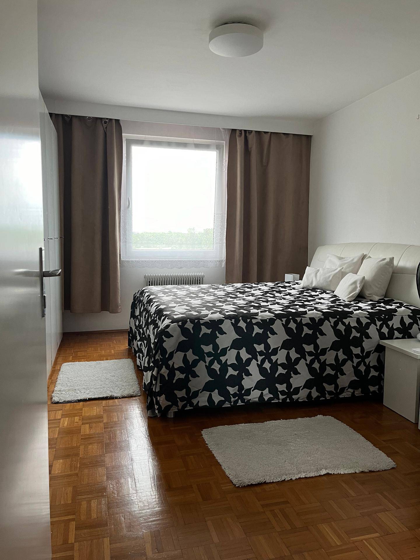 Schlafzimmer mit Parkettboden, großem Bett und Fenster mit braunen Vorhängen für Privatsphäre.
