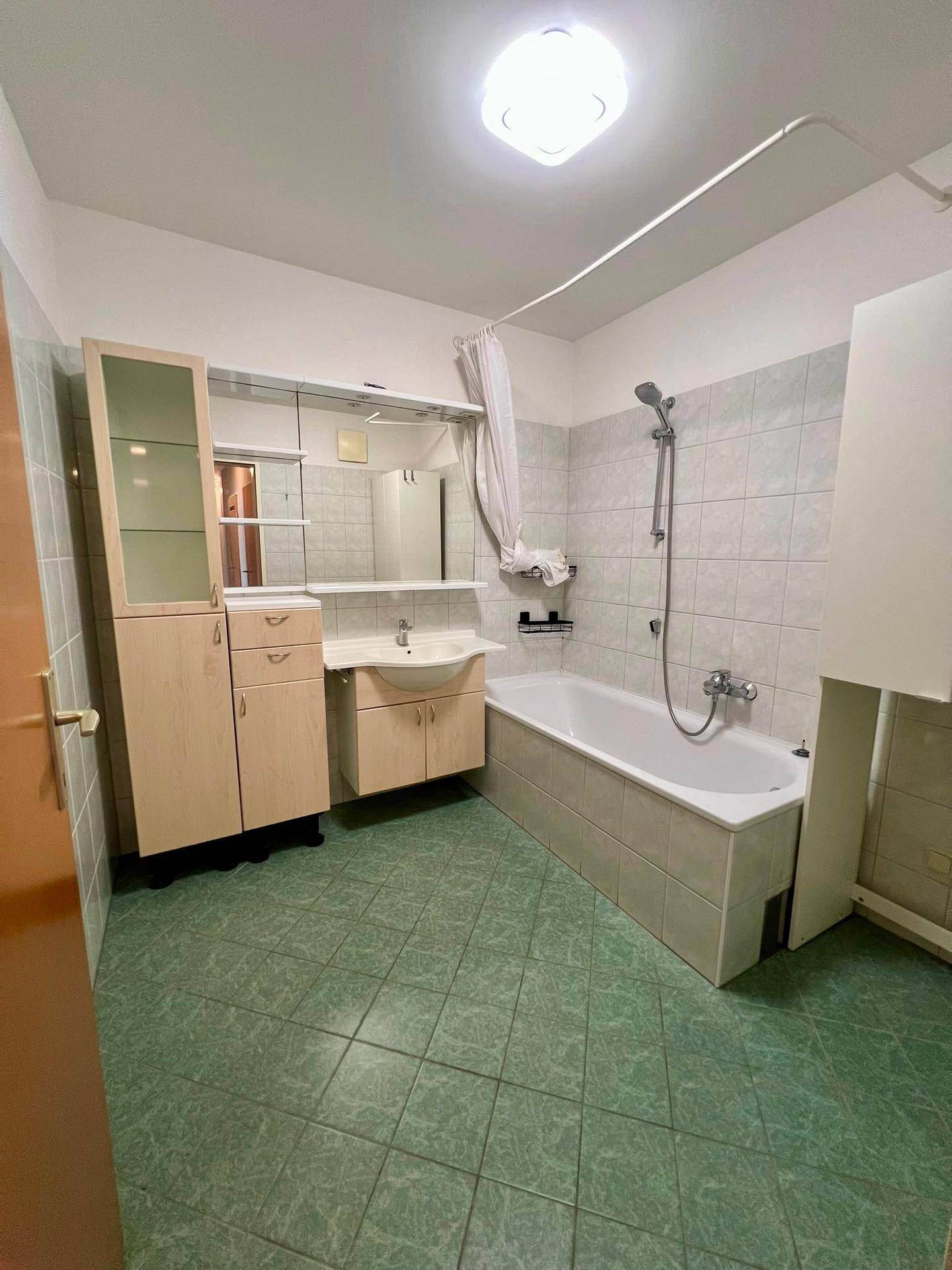 Badezimmer mit Badewanne, Duschvorhang, Waschbecken und grünen Bodenfliesen, funktional ausgestattet.