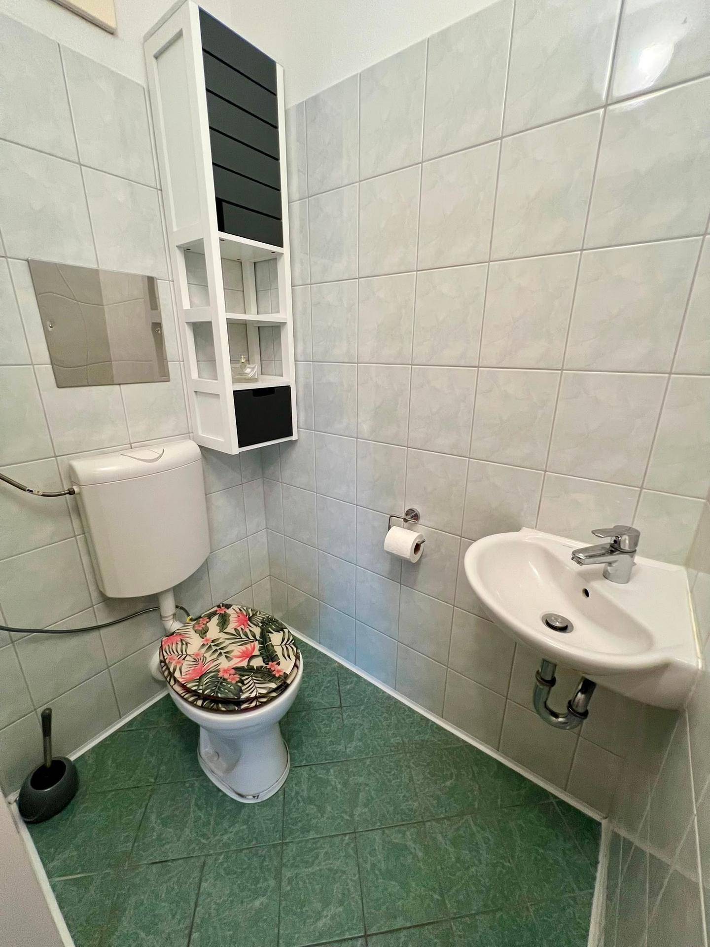 Kleines Gäste-WC mit grünen Fliesen, Eckwaschbecken und Wandregal, funktional eingerichtet.