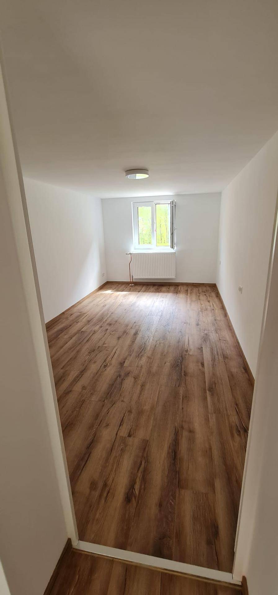 Leeres Zimmer mit hellem Holzboden, weißen Wänden und einem Fenster, das viel Tageslicht hereinlässt.