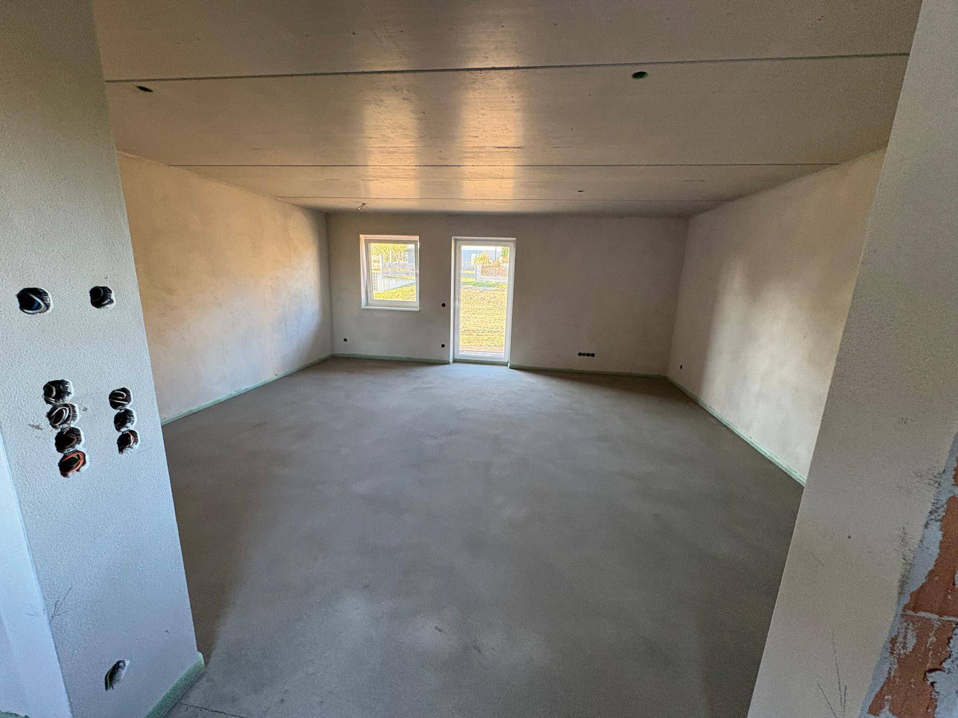 Geräumiger, unmöblierter Wohnbereich im Neubau mit mehreren Fenstern und Zugang nach draußen.